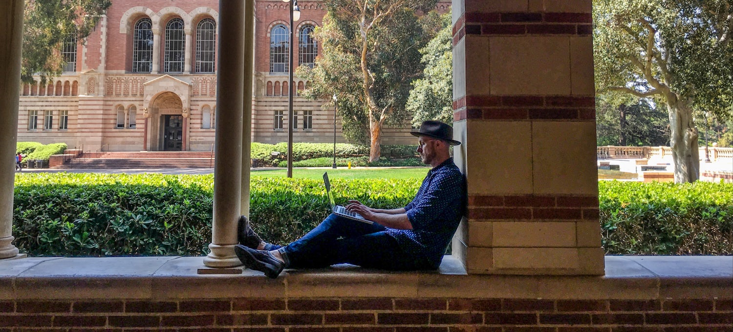 [Featured image] A man working on getting his master's degree studies on a laptop outside on a university campus.