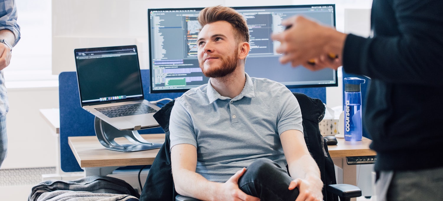 [Featured Image] A machine learning engineer in a gray shirt sits with his back to his desk and speaks with co-workers.