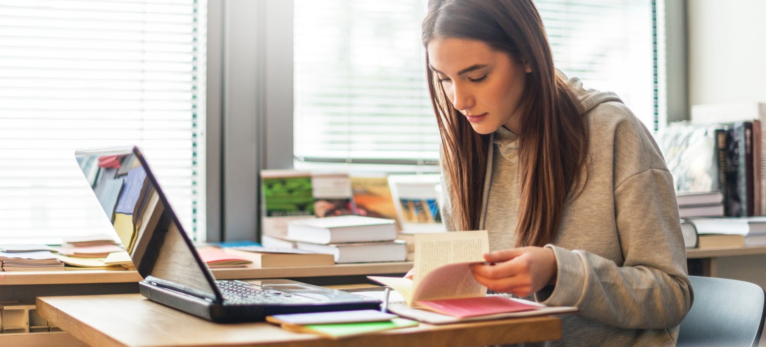 [Imagen destacada] Una estudiante de licenciatura en un escritorio con su ordenador portátil y un libro abierto.