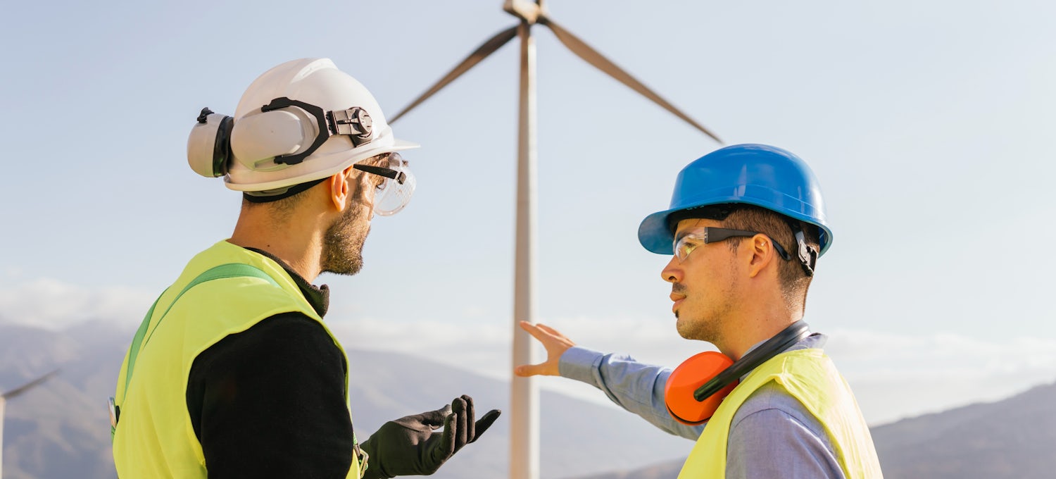[Featured Image] A renewable energy engineer talks to a technician at a wind turbine.  