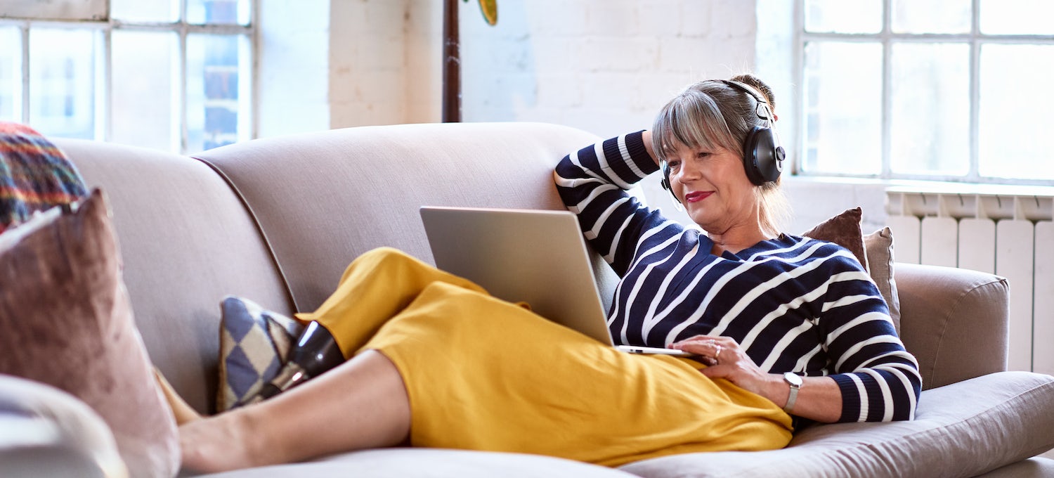 Woman with gray hair, a navy and white striped sweater, and a yellow skirt slightly smiling while lounging on a couch attending online school on her laptop.