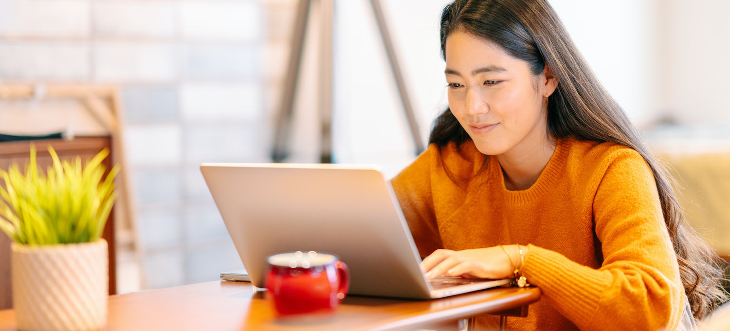 [Featured image] A cloud engineer wearing a bright orange sweater works at her laptop with a hot beverage in a bright office or cafe.