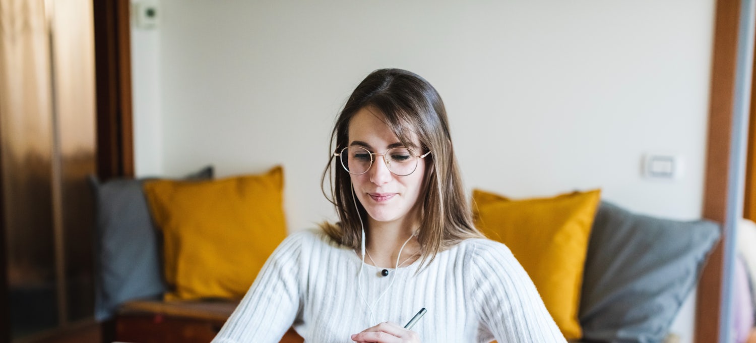 [Featured Image]:  A female wearing a white top and glasses. She has long brown hair. She is sitting in front of her laptop, holding a pen in her hand. 