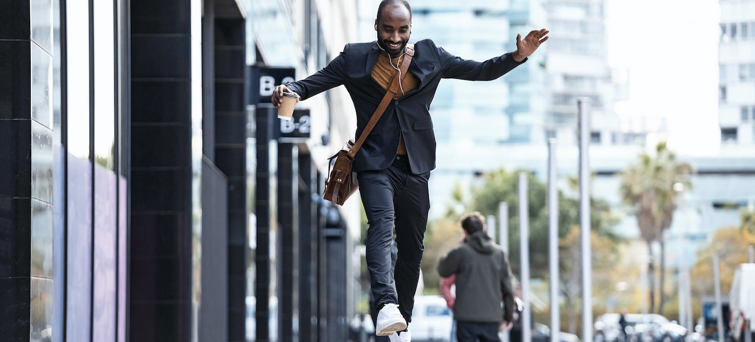 A smiling professional wearing a shoulder bag and carrying a to-go cup of coffee leaps from one bench to the next outside an office building.