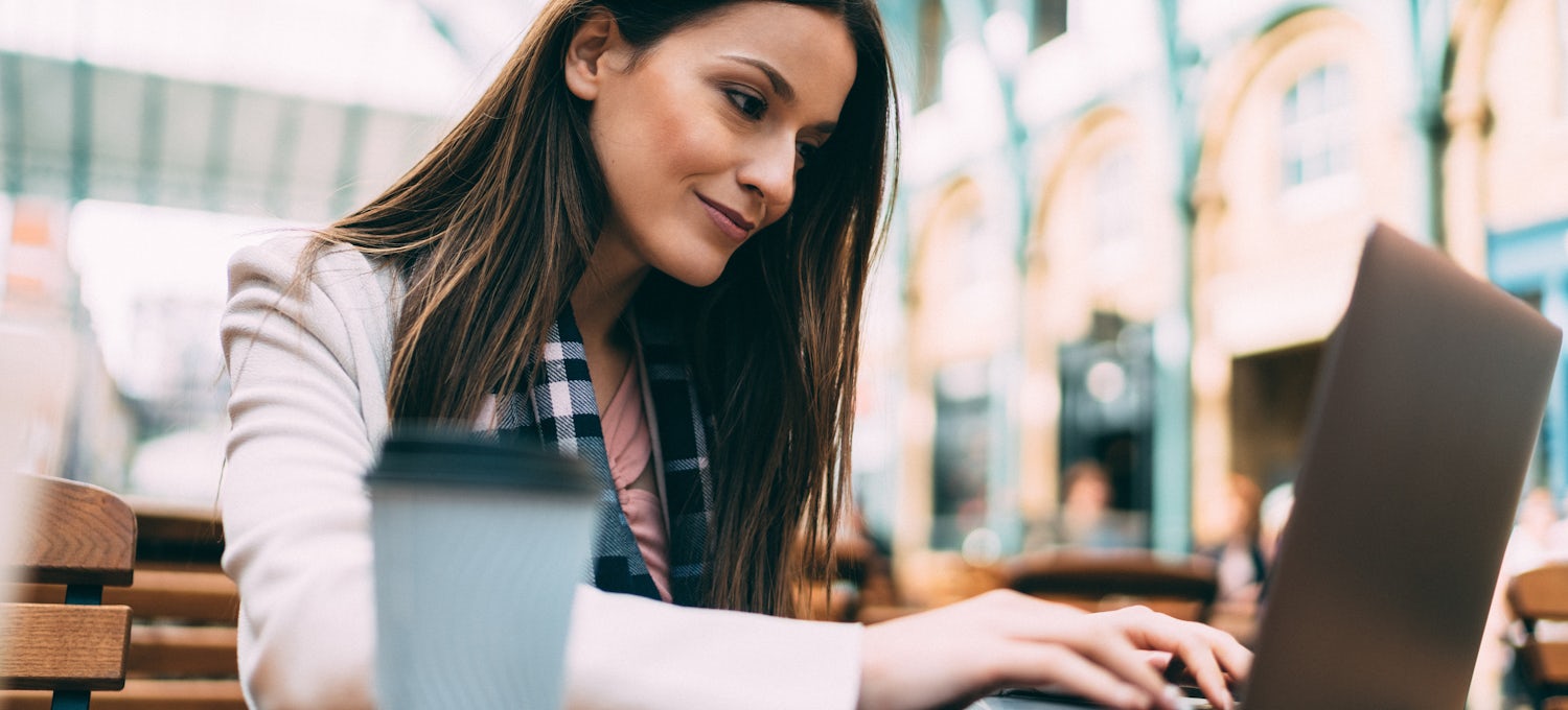[Featured Image] A penetration tester sits outdoors with a cup of coffee and uses her laptop to look for potential cybersecurity financial sector jobs. 
