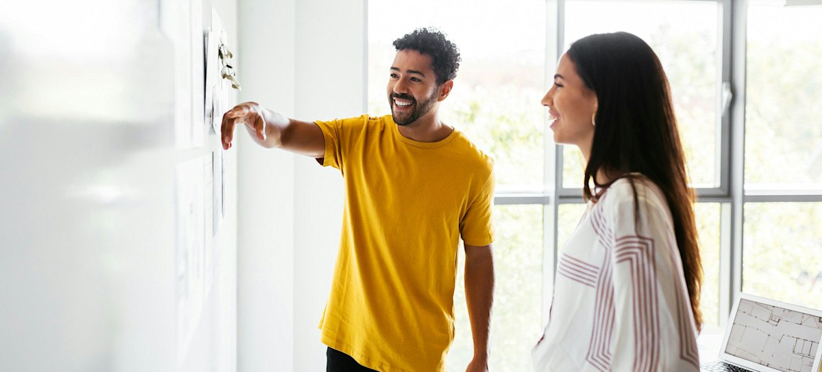 [Featured Image] Two coworkers review materials hung on a wall containing an AI-supported marketing plan. 