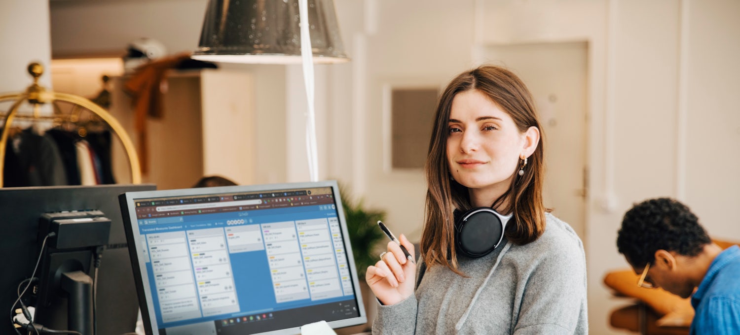 [Featured Image]  A person in a gray sweater with headphones around their neck sits at a shared desk and reviews project management software on a monitor to address scope creep.