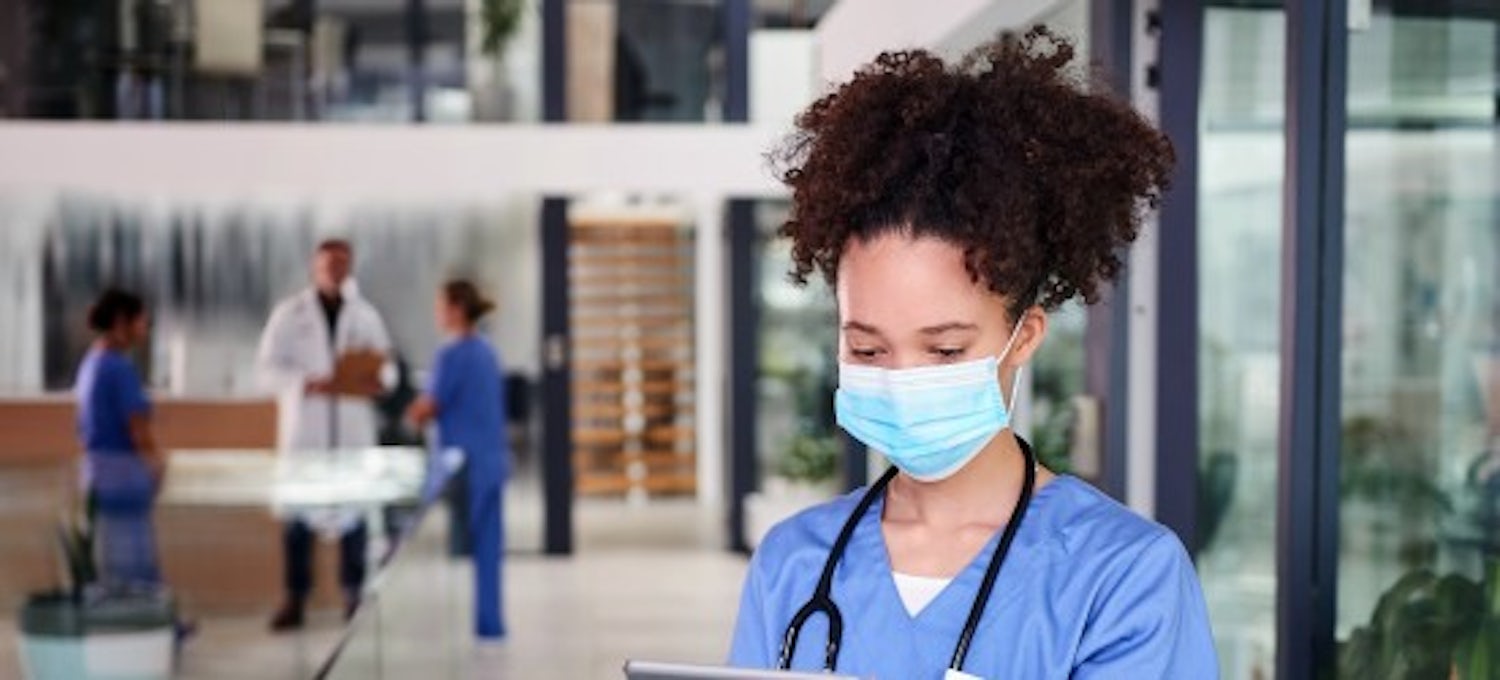 [Featured Image]:  A female, with black hair, wearing a blue uniform, face covering, and a stethoscope around her neck is looking at a chart standing in a medical office.