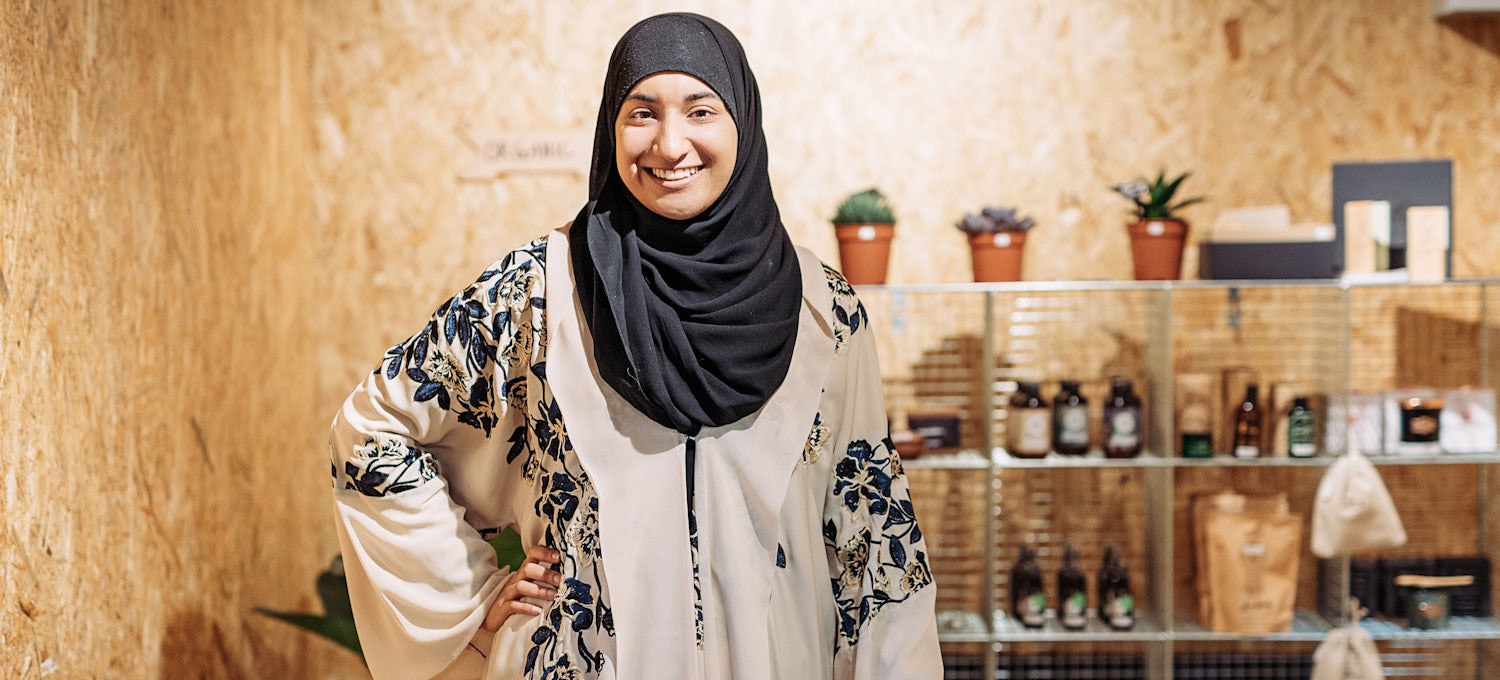 [Featured Image] A woman in hijab smiles and stands before the products she sells through her home business. 