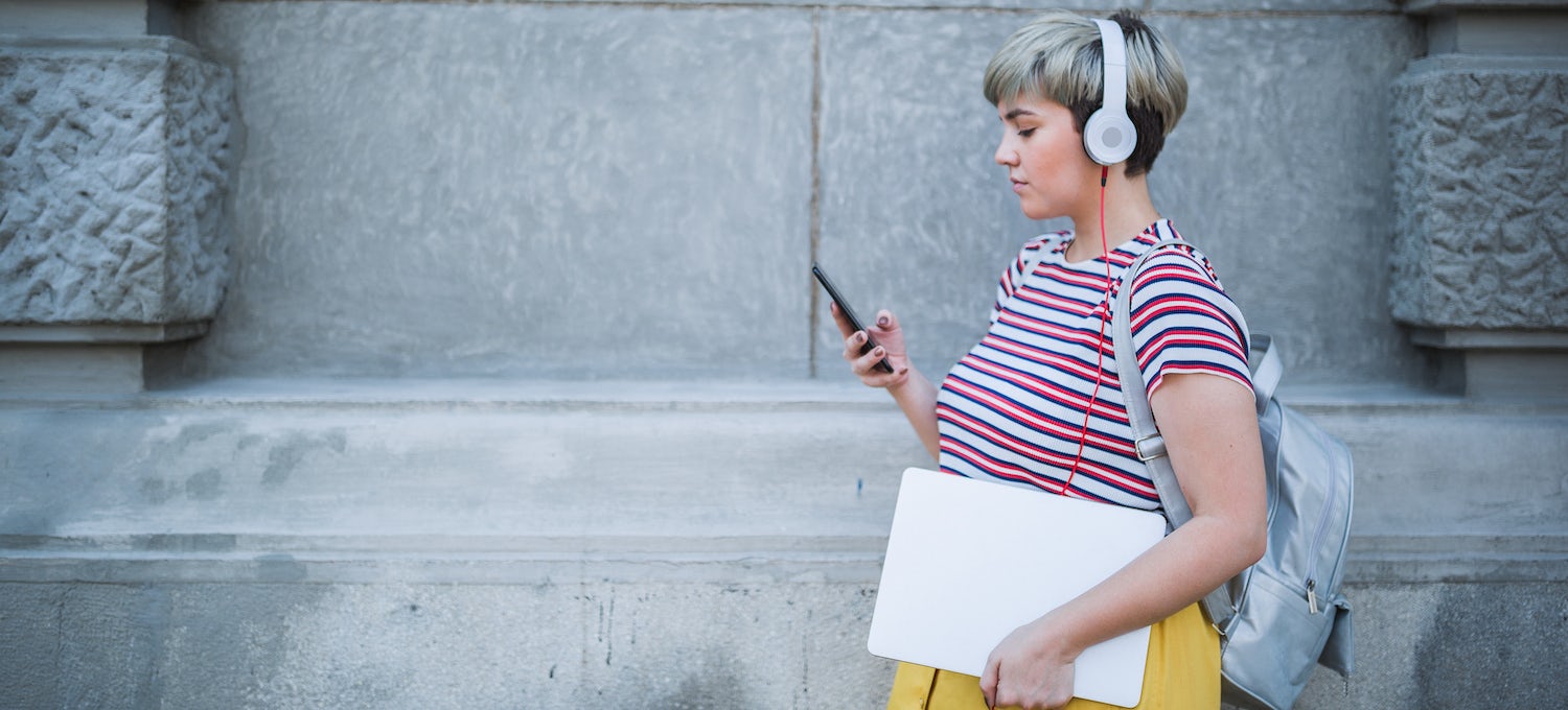 [Featured image] A young person wearing over-the-ear headphones and carrying a notebook looks at their phone. 