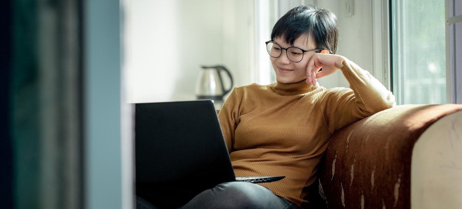 [Featured image] A young Asian woman with short hair and glasses sits on her couch smiling into her laptop.
