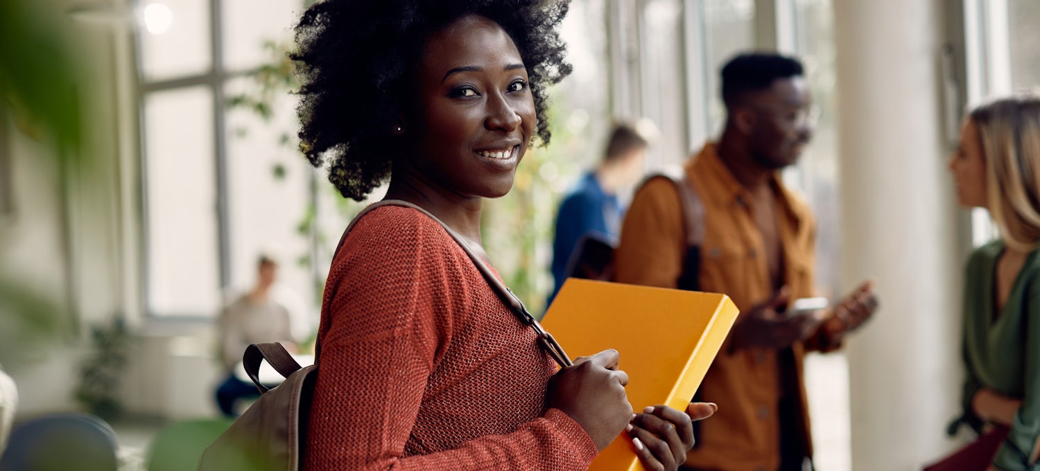 [Featured image] A business degree student in a coral sweater with a backpack on stands in a hallway with a yellow binder in her arms.