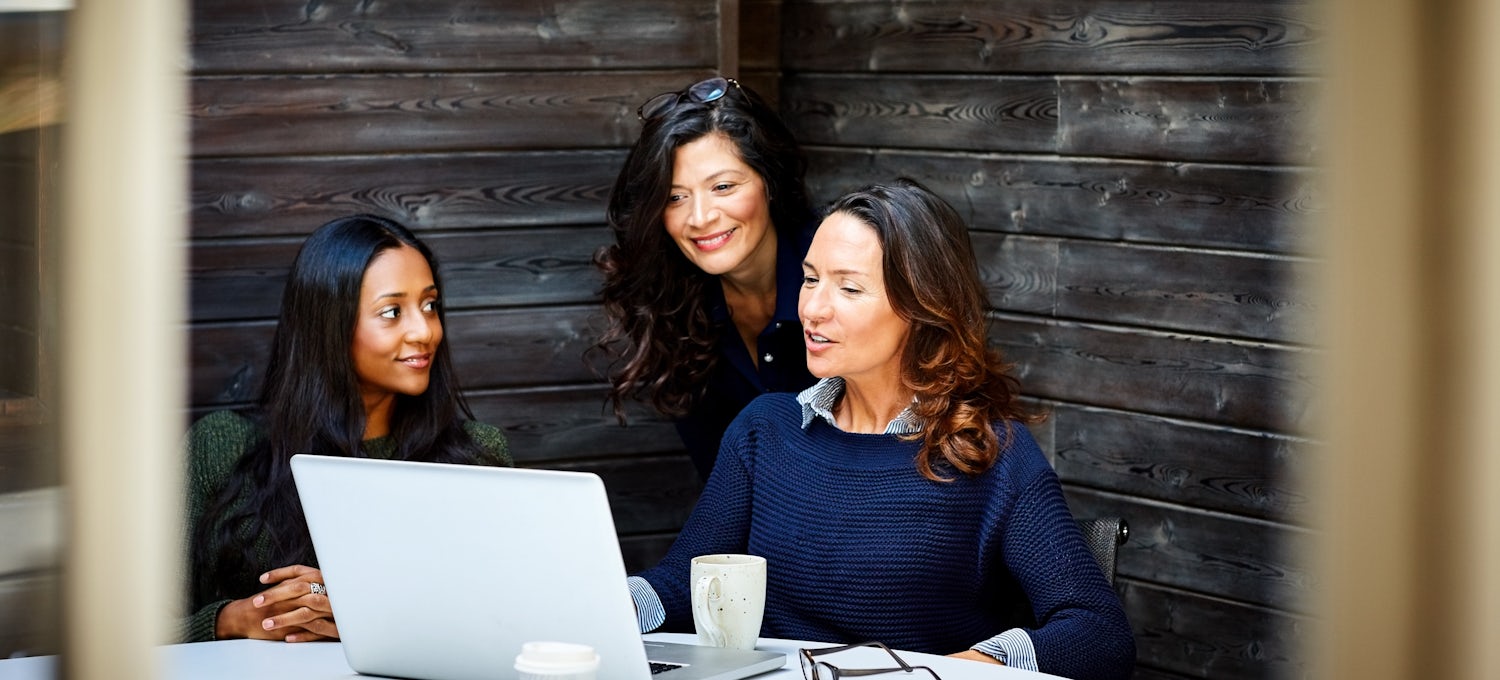 [Featured image] A business woman with brown hair and blue sweater chats with two female colleagues about a proposal on her laptop.
