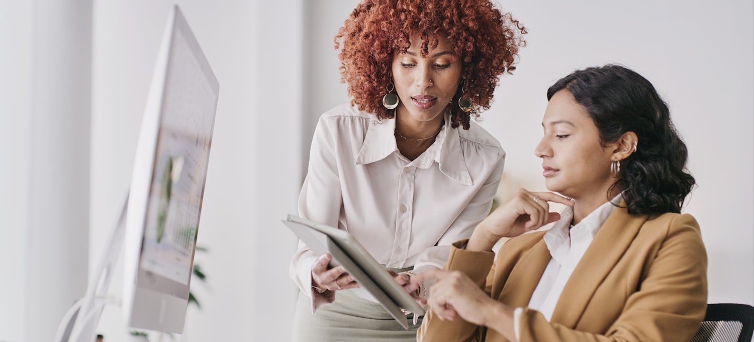 [Featured image] An employee approaches a member of middle management with a question about some information on her tablet.  
