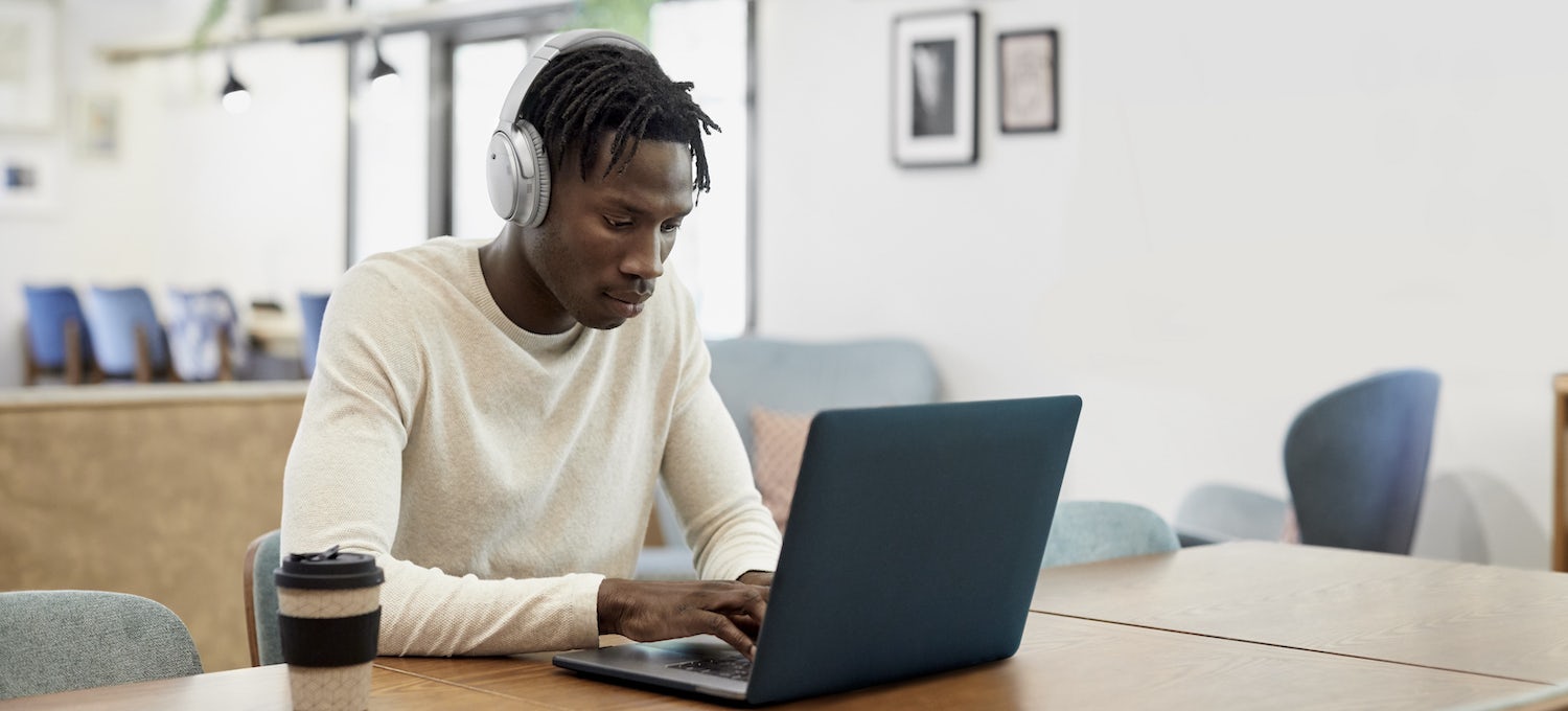[Featured Image] A PhD student works on his laptop while listening to over-the-ear headphones. 