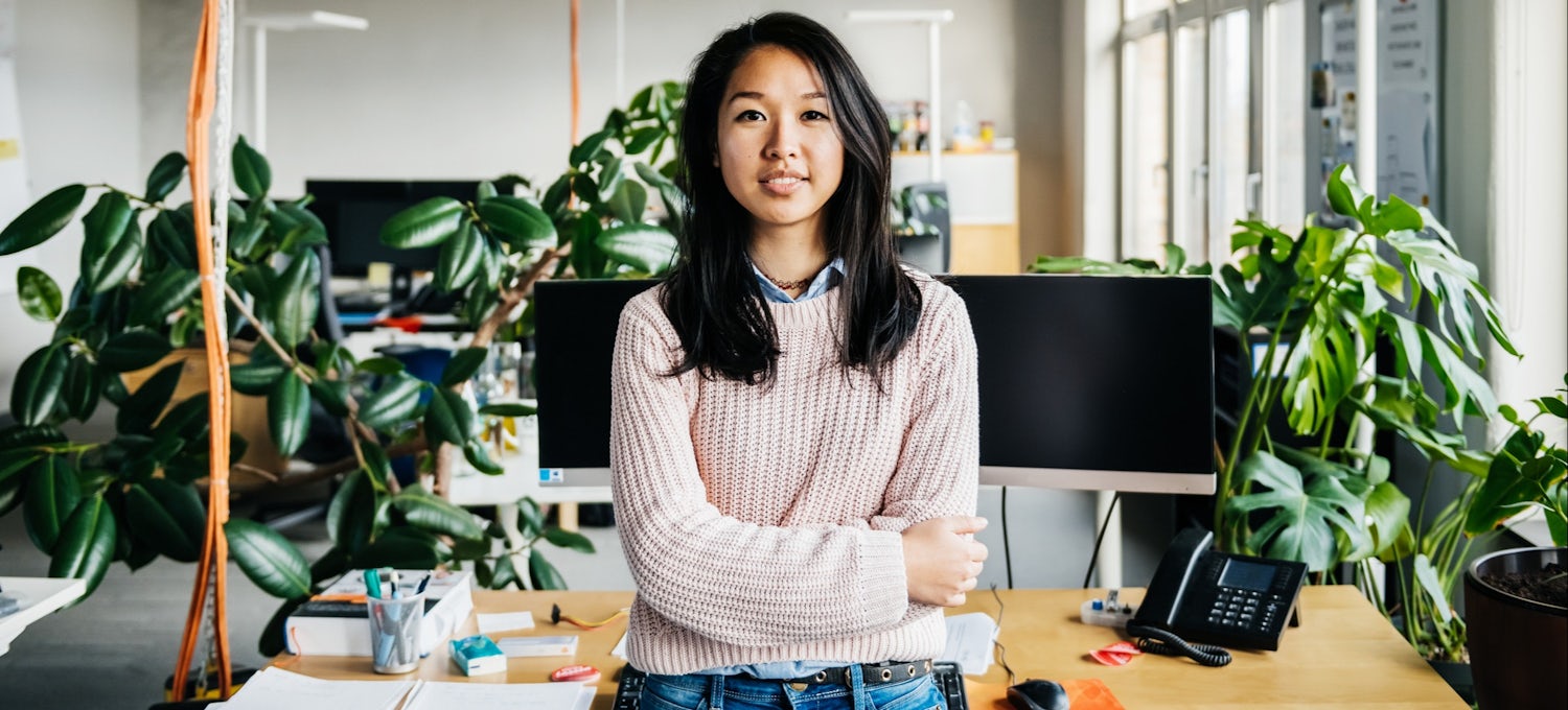 [Featured image] A DevOps engineer in jeans and a pink knit sweater stands in front of her desk and smiles at the camera. Her workspace has two monitors, a telephone, and several indoor plants.