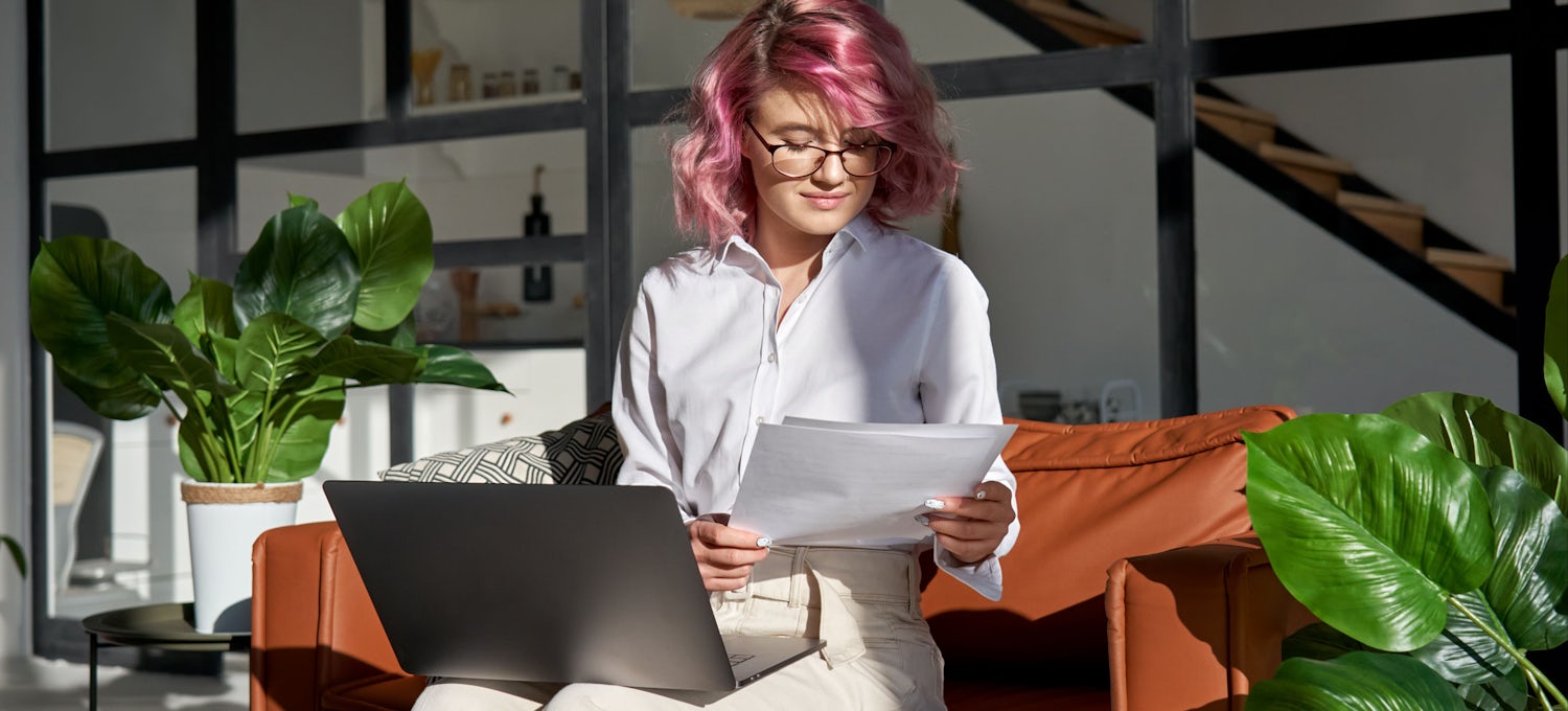 [Featured Image] A lady with pink hair is holding a piece of paper with a laptop on her lap.
