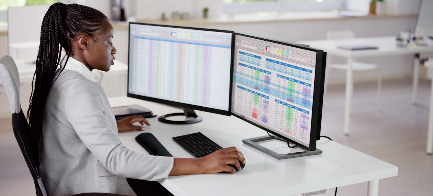 [Featured Image] A data analyst sits at her office desk analyzing spreadsheets on her two computer screens to gain data insights about her work.
