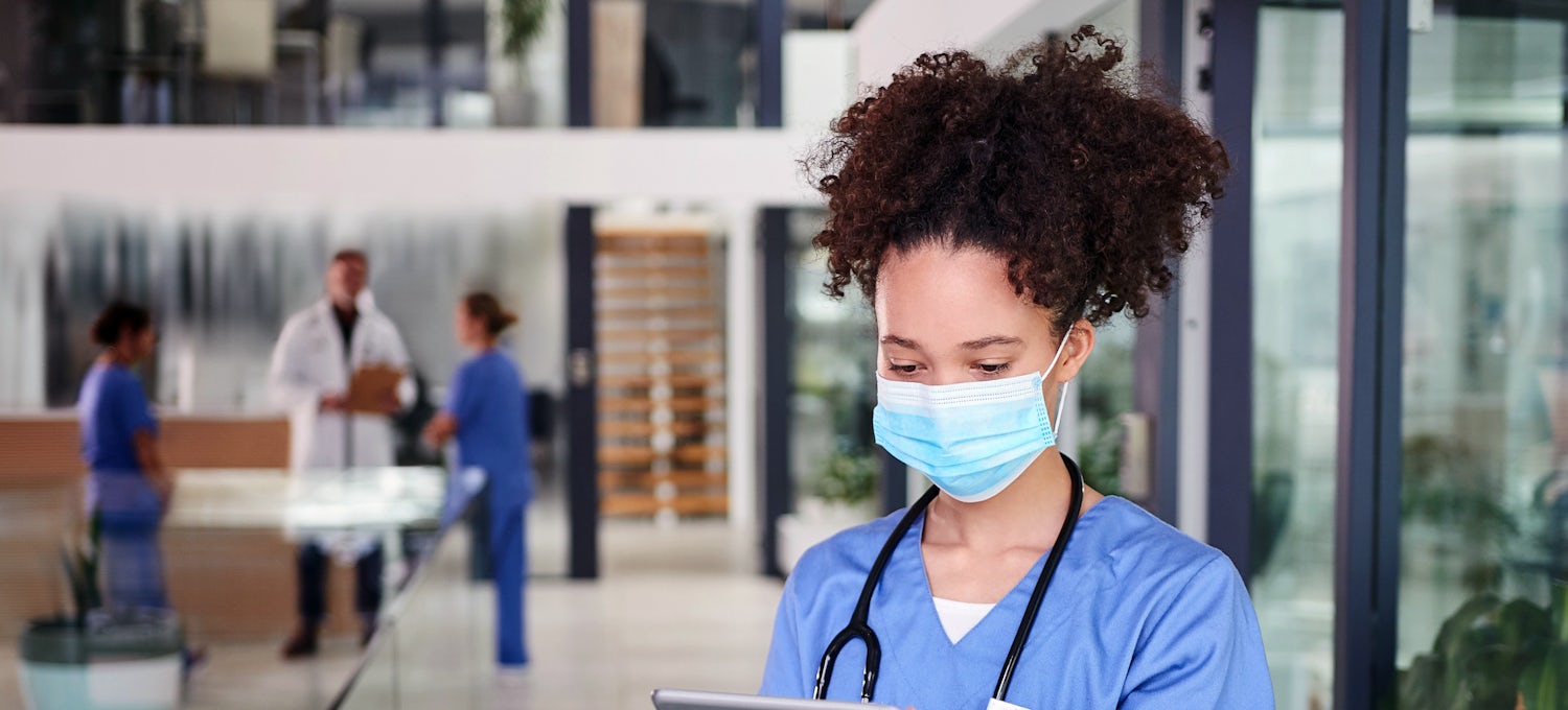 [Featured Image]: Nurse anesthetist, wearing a blue uniform and blue mask, standing in exam area, reading charts of a patient.