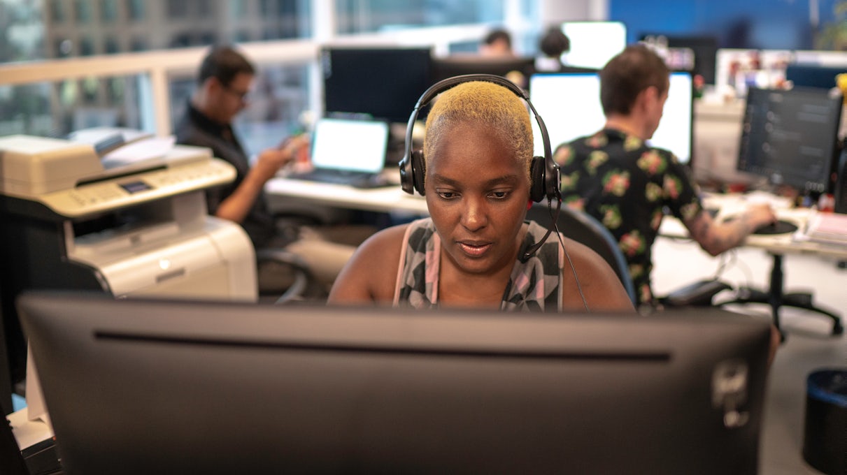 [Featured image] An information security analyst wearing black headphones and a patterned tank top works at a monitor with a printer to their right and colleagues in the background.