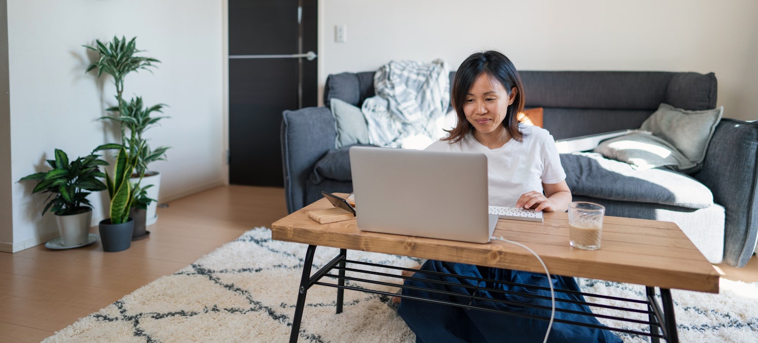 [Featured Image] A woman in a white t-shirt sits on the floor at her coffee table and types prompts into ChatGPT for a project.