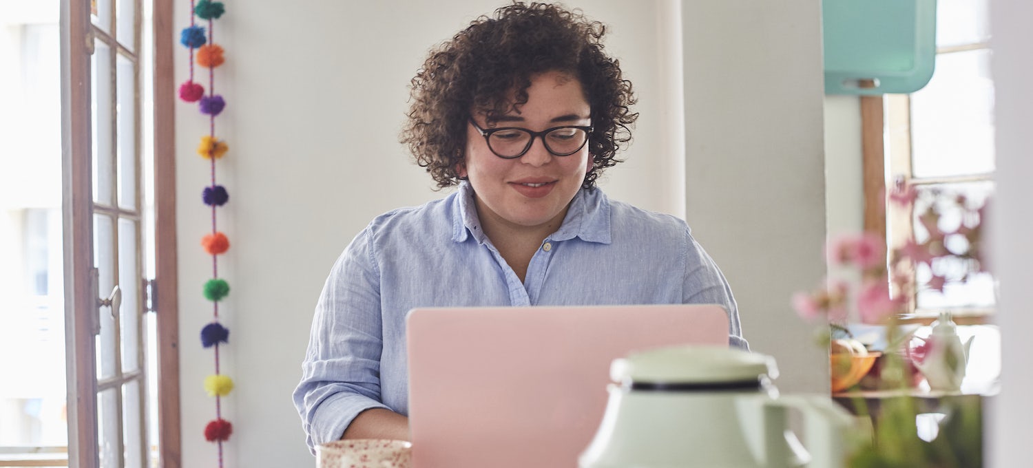 [Featured image] A woman with curly hair and glasses looks at her pink laptop at home.