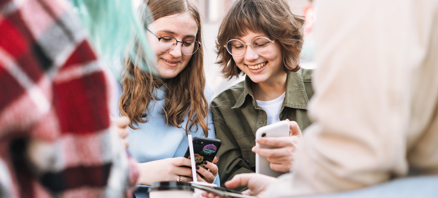 [Featured image] Two girls at a table outside in the city looking at their phones and talking about going to college after earning their GED.