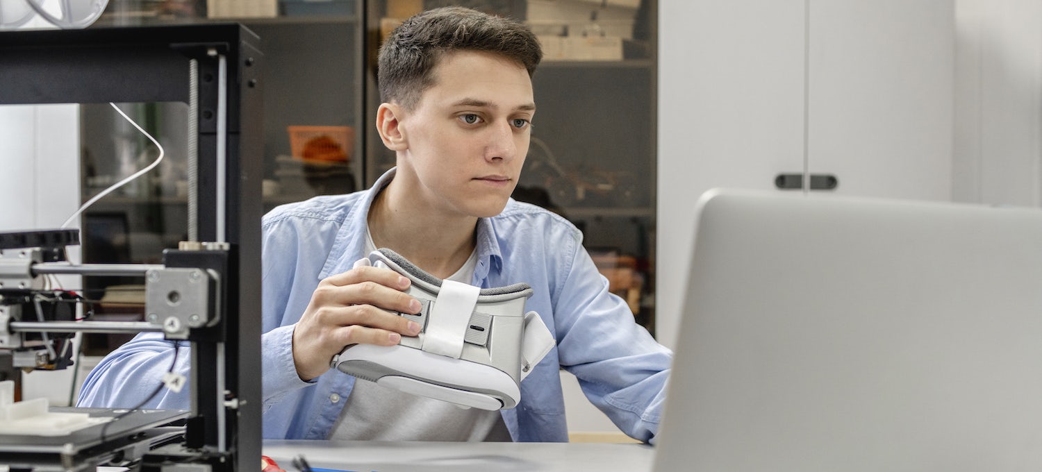 [Feaured image] A young white man looks at his laptop while holding a VR headset. 