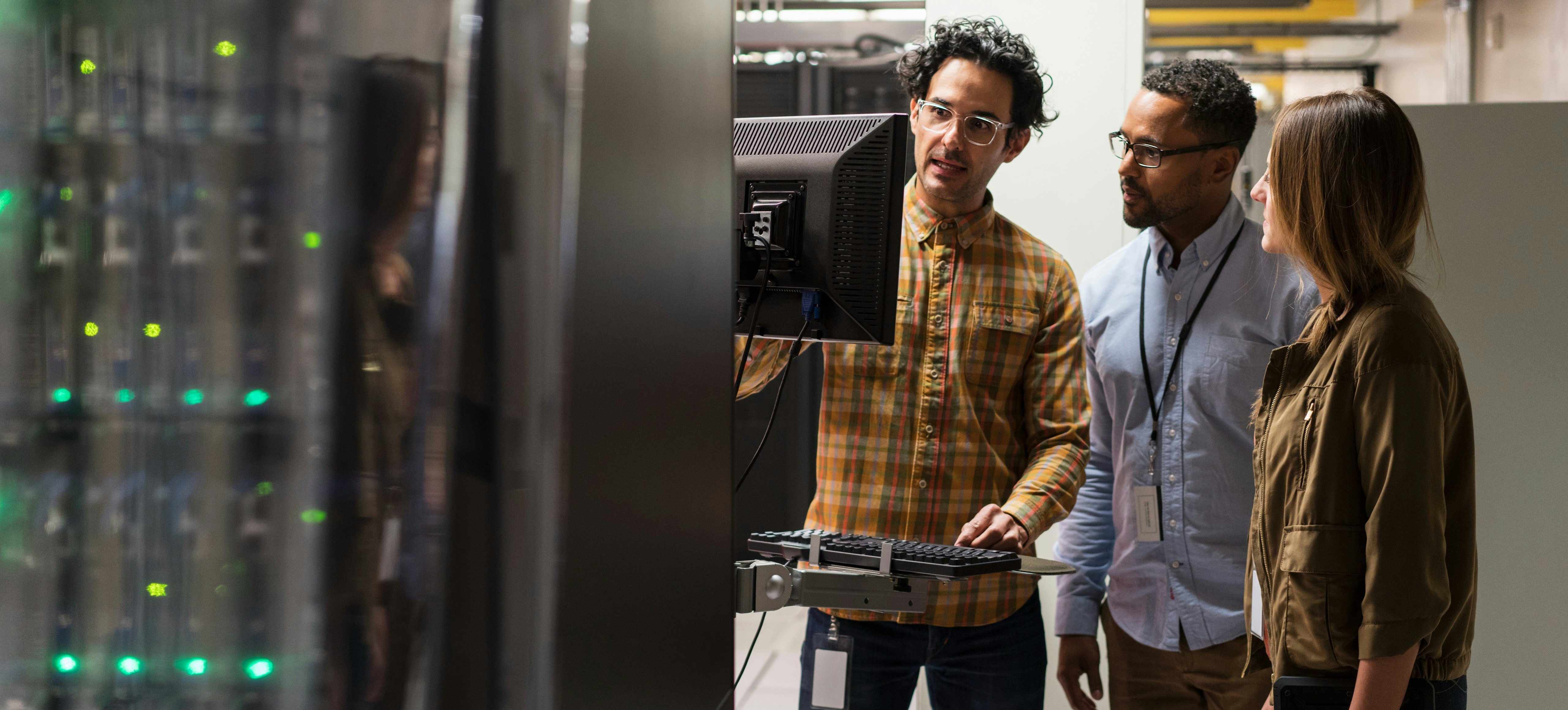 [Featured Image] A network administration answers, "What does the term attenuation mean in data communication?" as they talk to two coworkers gathered around a desktop computer station in a networking room.