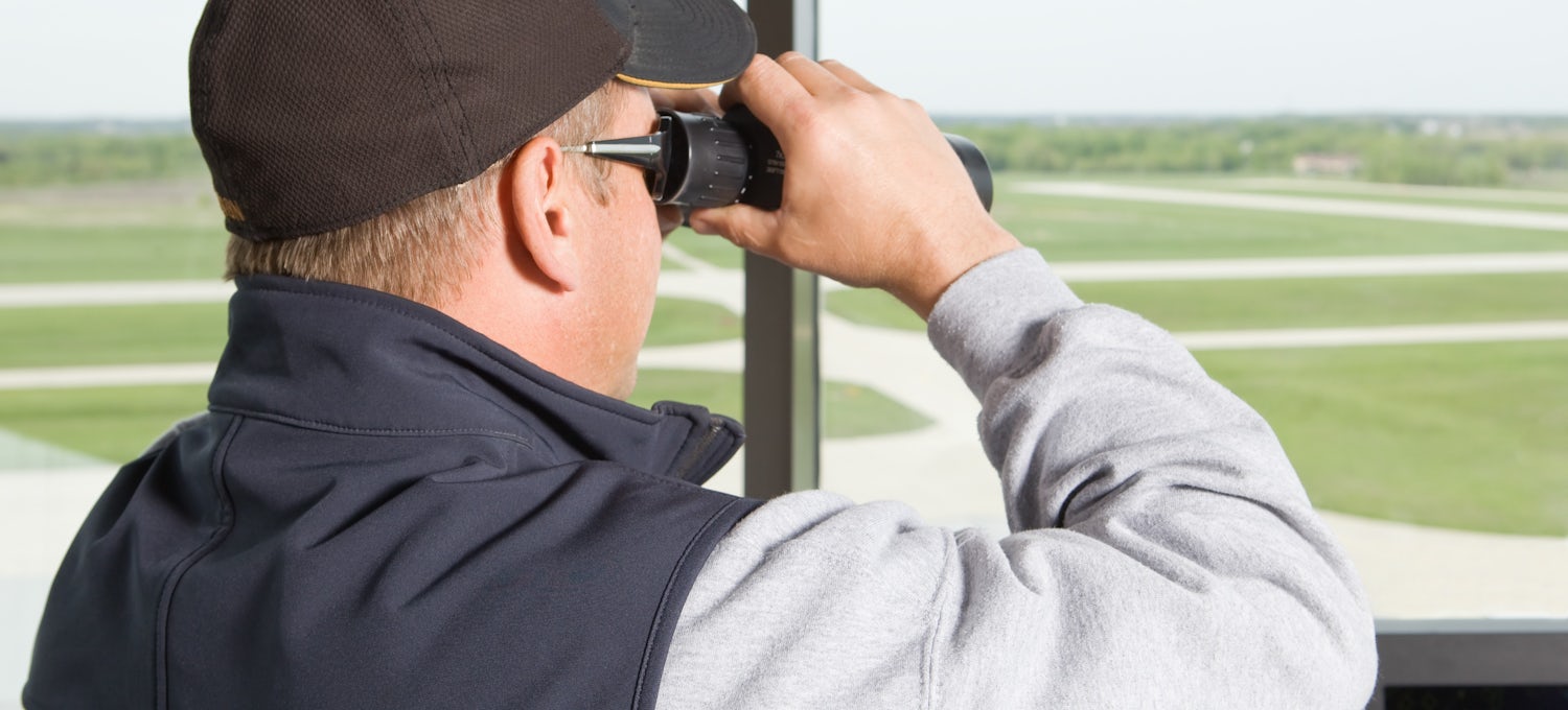 [Featured Image] An air traffic controller, which is one of many jobs that pay $50 an hour without a degree, uses binoculars to look out a window over a runway.
