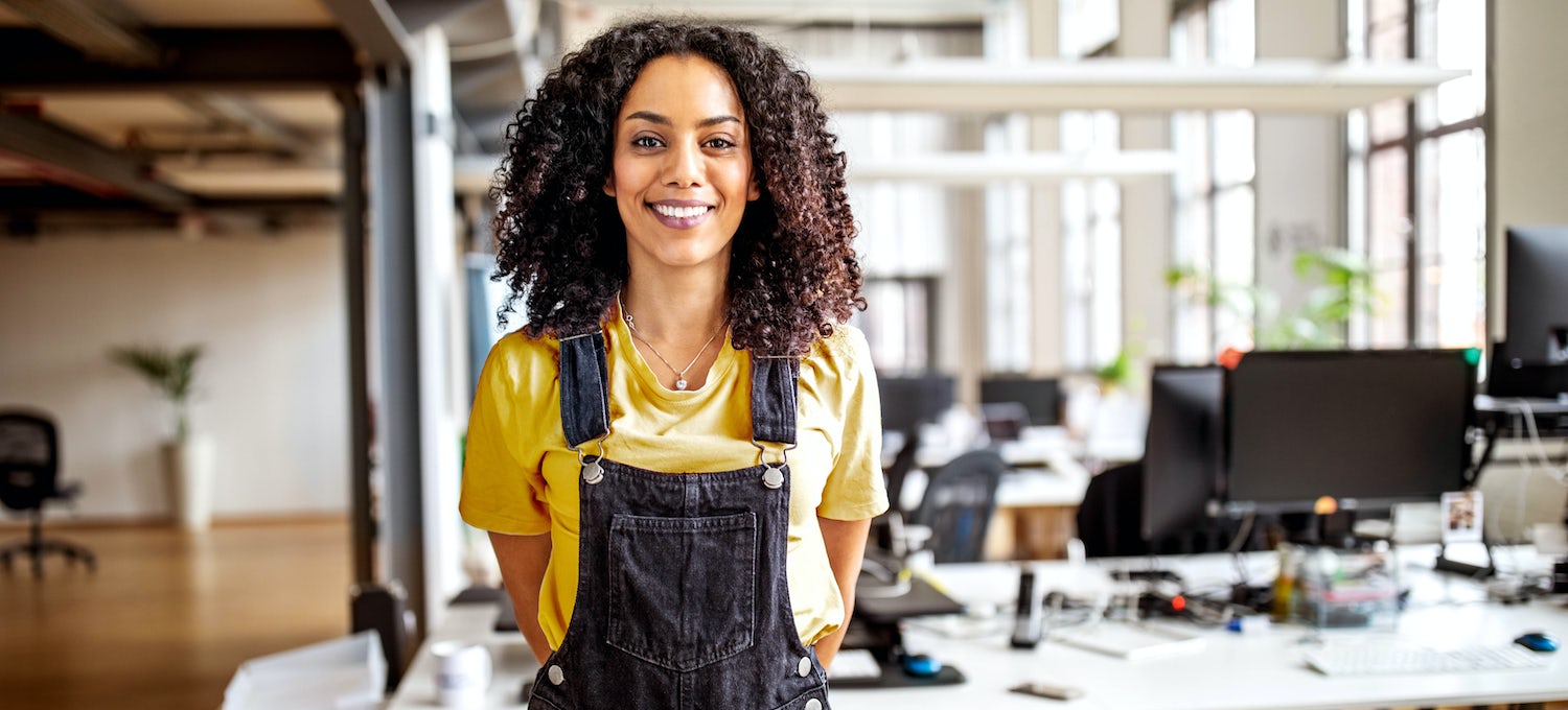 [Featured image] A solutions architect wearing black overalls and a yellow shirt stands alone in an open office.