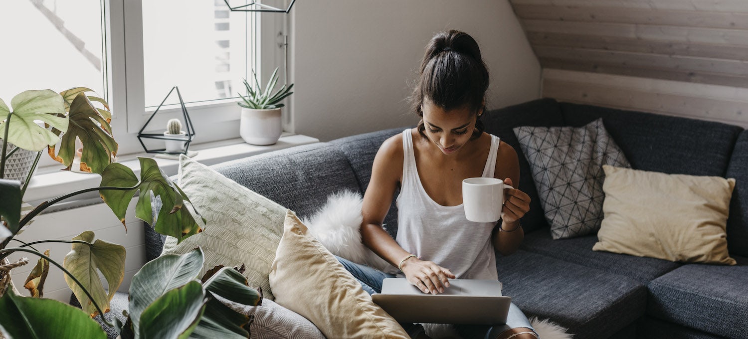 [Featured image] A woman with her hair in a ponytail holds a cup of coffee while using her laptop on the couch.
