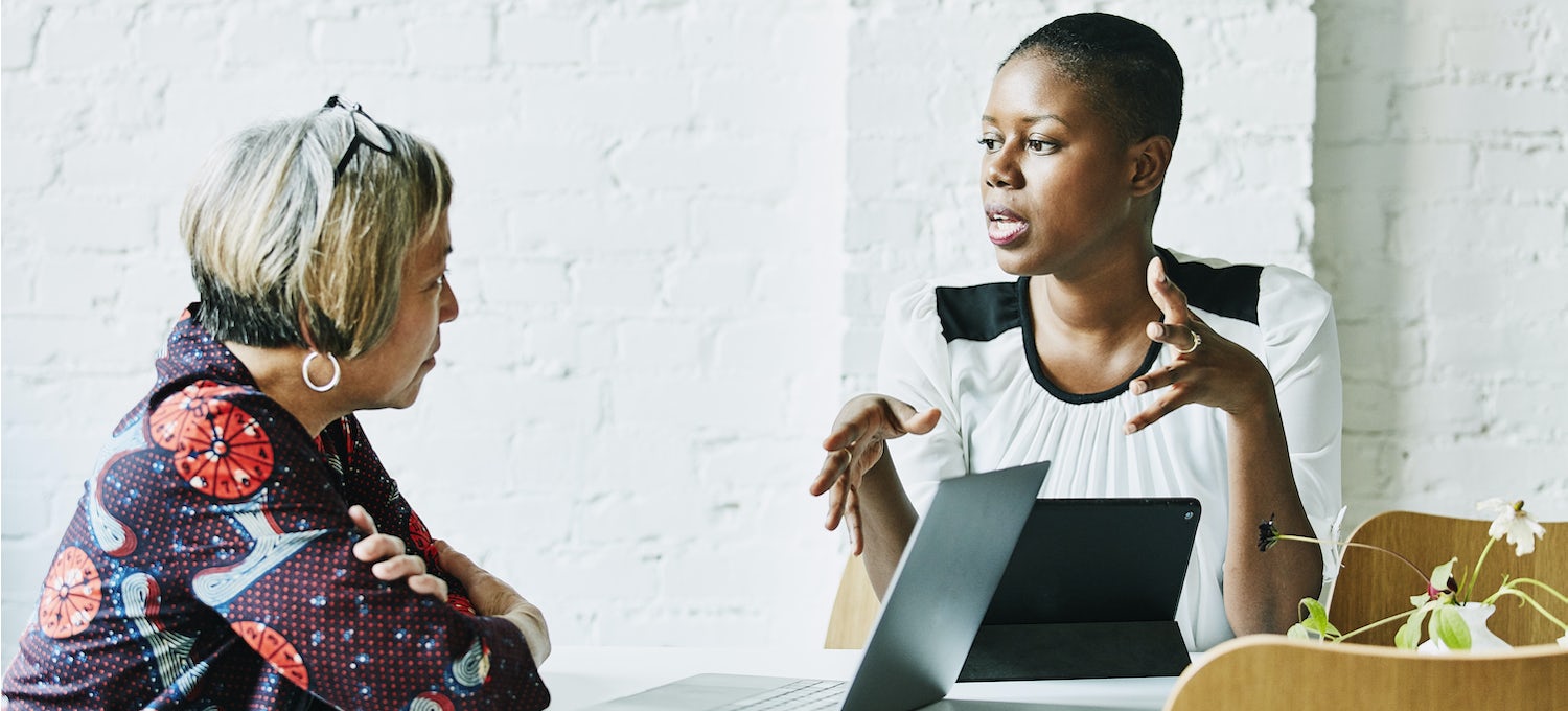 [Featured image] A female business analyst interviews a female employee. Both have their laptops in front of them.