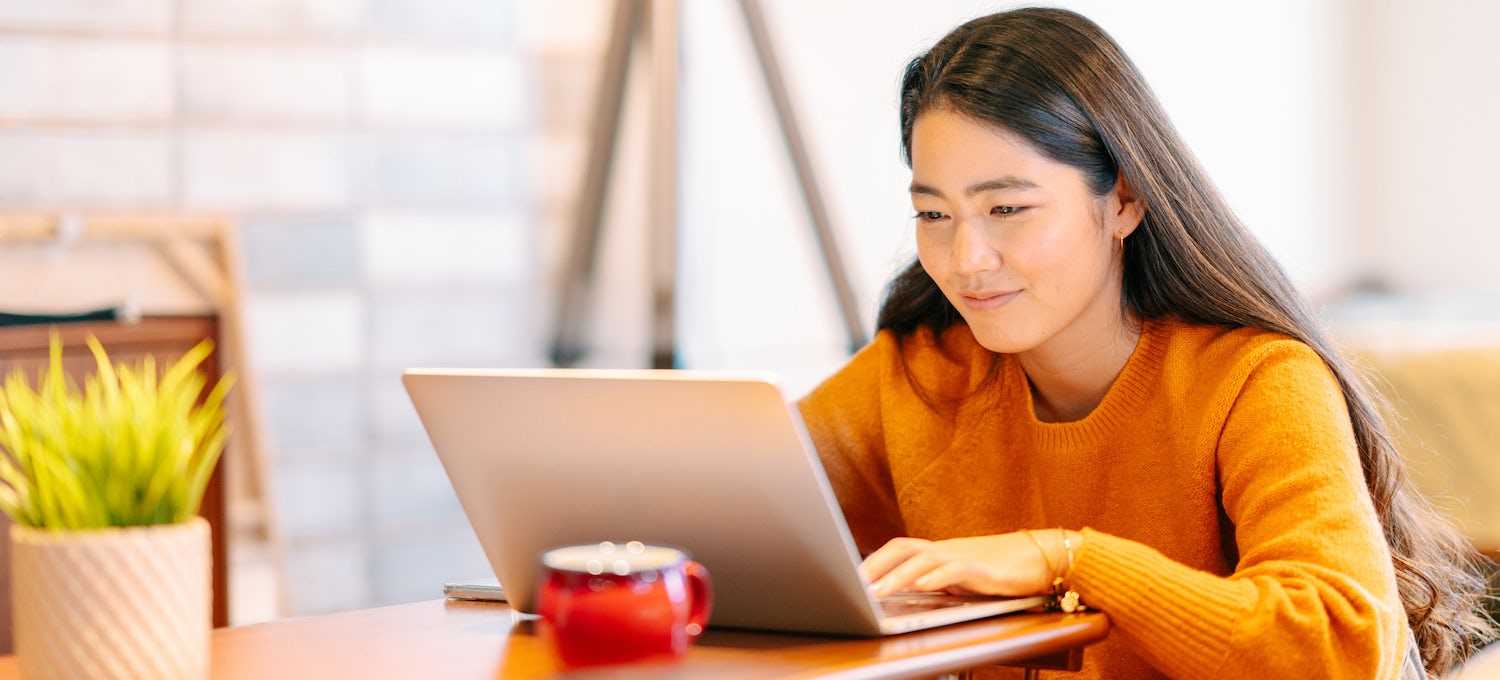 [Featured image] A young woman wearing an orange sweater looks at her laptop. 