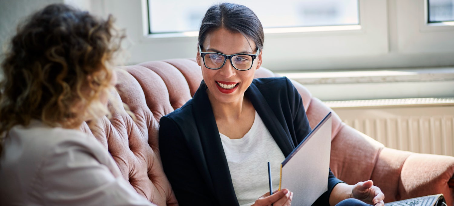 [Featured Image] Woman in glasses interviewing a second woman while sitting on a couch in an office