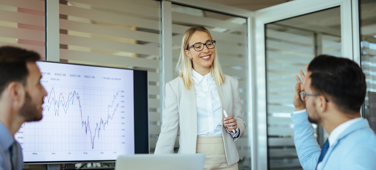 [Featured image] A female, wearing a white business casual outfit and glasses, is leading a meeting with the marketing team, two males, in her office. She is standing in front of her laptop and a screen showing a graph of the effects of inbound marketing.  