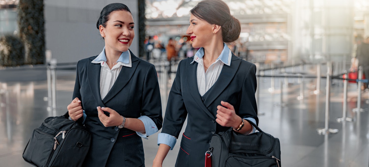[Featured Image] Two flight attendants carry their bags and talk while walking through an airport.
