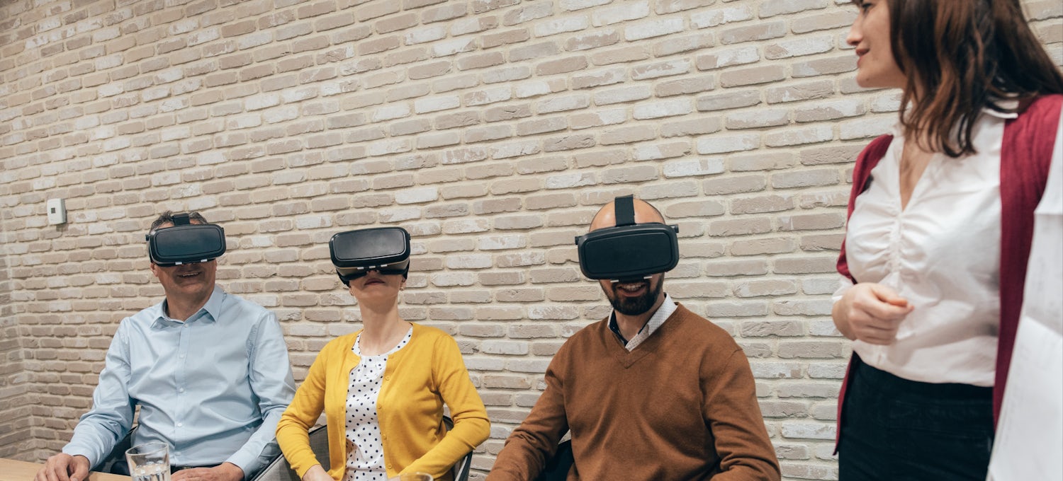 [Featured Image] A group of colleagues sit in a business meeting wearing virtual reality headsets and experimenting with VR, one of the top business intelligence trends of 2024. 
