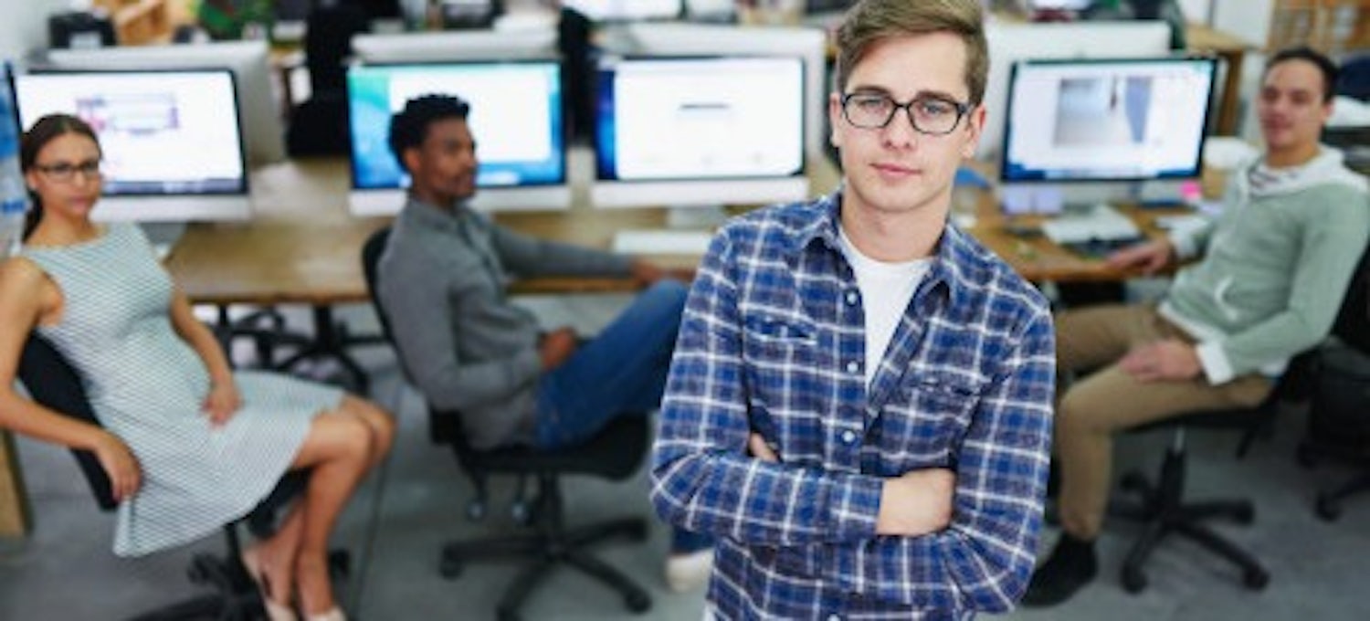 [Featured Image] A DevOps engineer in a blue button-down shirt stands in front of three members of his development team.