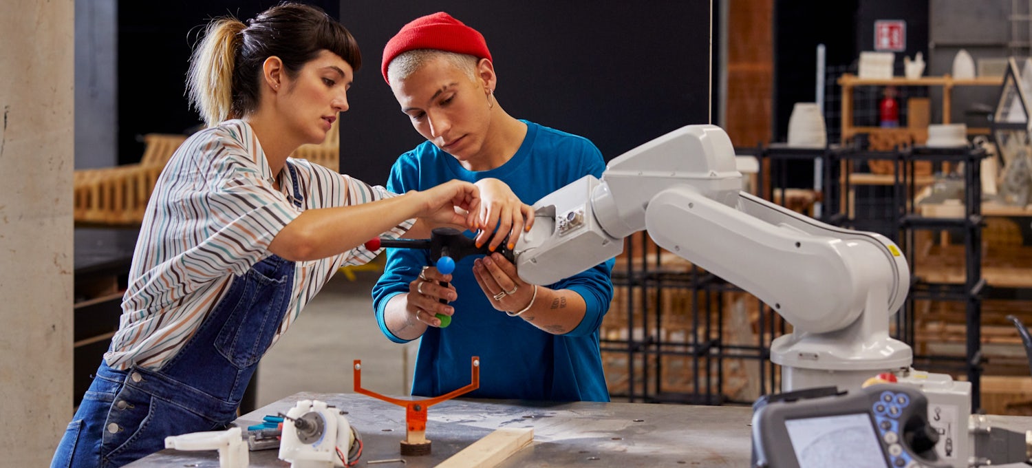 [Featured Image] Two robotics engineers work together on a robotic arm in a warehouse.
