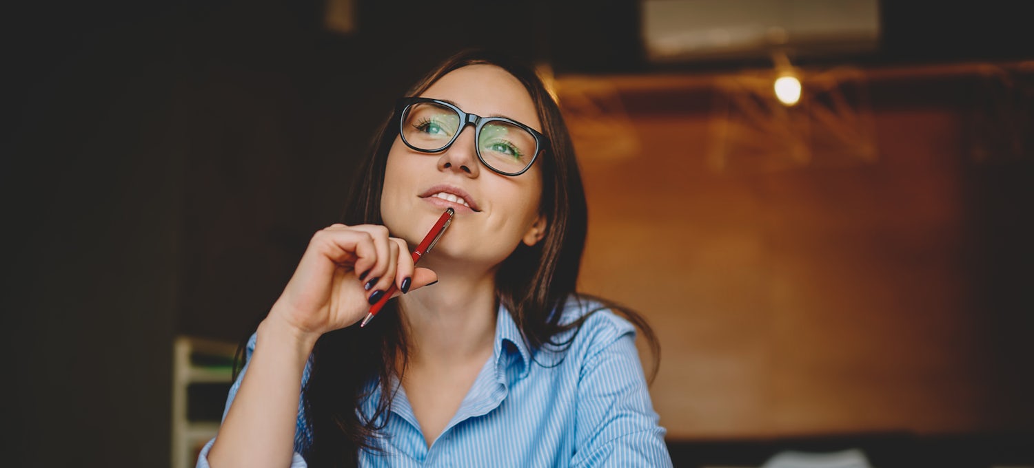 [Featured Image]:  A woman with long brown hair, wearing a blue blouse and glasses, and a watch is holding a pen in her hand, up against her chin. She is thinking with open books in front of her. 