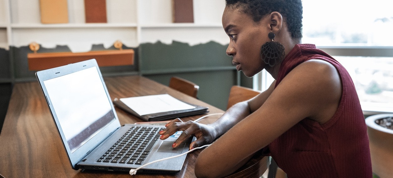 [Featured Image] A woman in a maroon shirt sits at a wooden table and works on her LinkedIn profile on a laptop. 