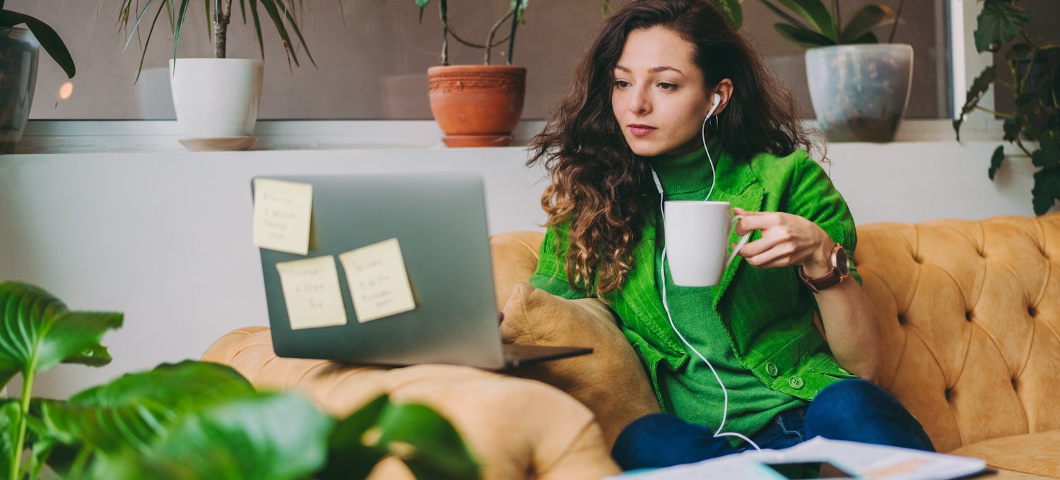 [Featured image] An email marketing manager in a green shirt and sweater sits on an orange sofa and works on her laptop. She's holding a white mug.