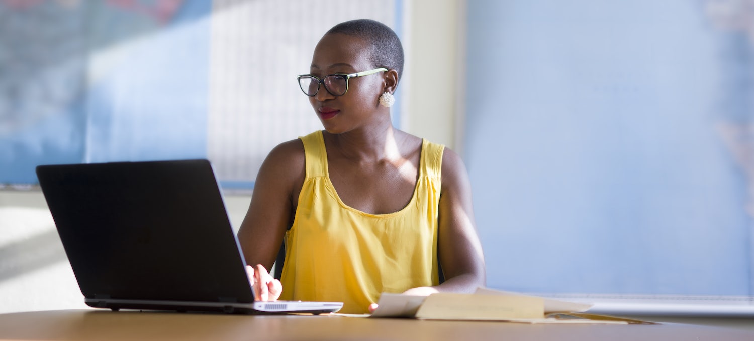 [Featured image] A data analyst wearing a yellow tank top and eyeglasses sits in front of a laptop in an office writing a statistical analysis program.