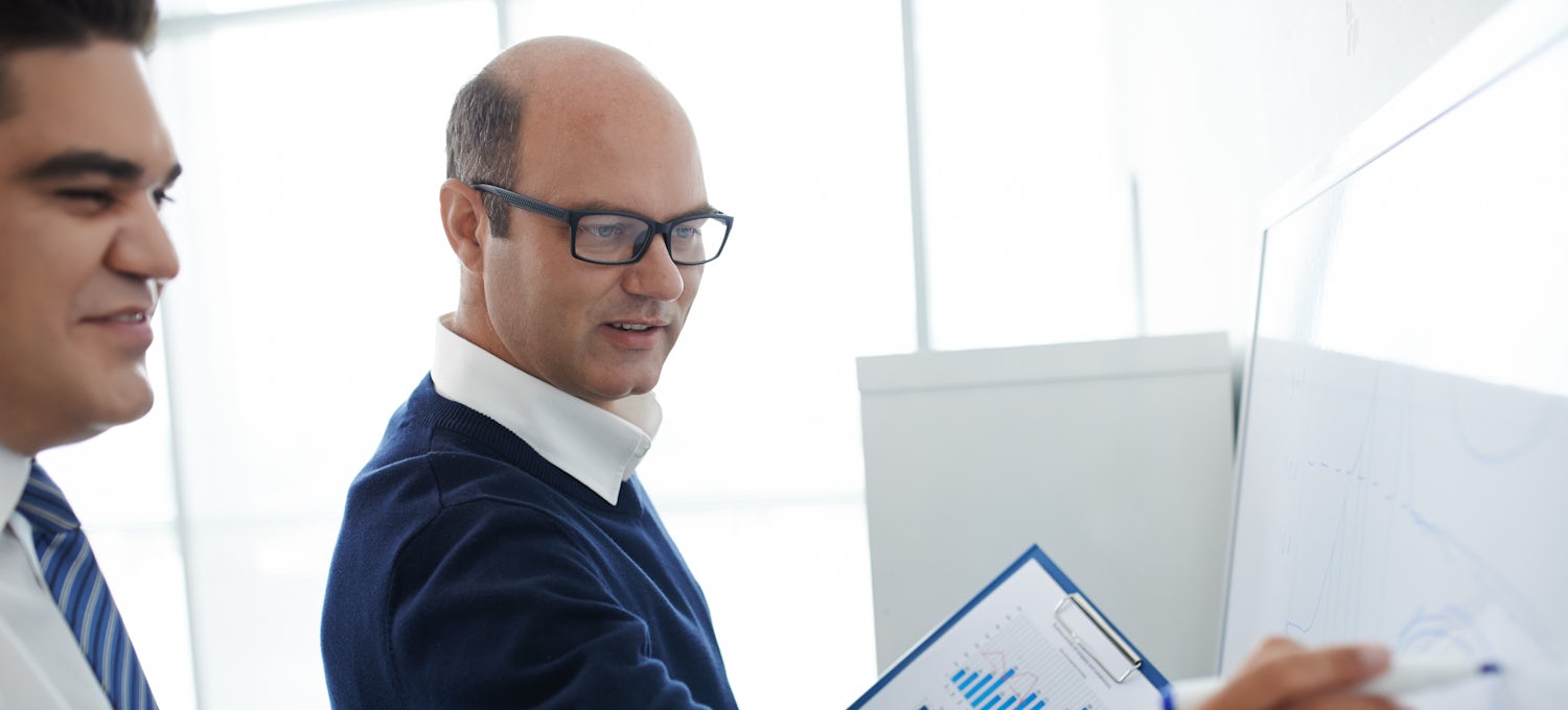 [Featured Image] A project manager, wearing a blue sweater and white shirt, is standing at a board and holding documents with graphs and tables, discussing the latest project strategy with his team member.
