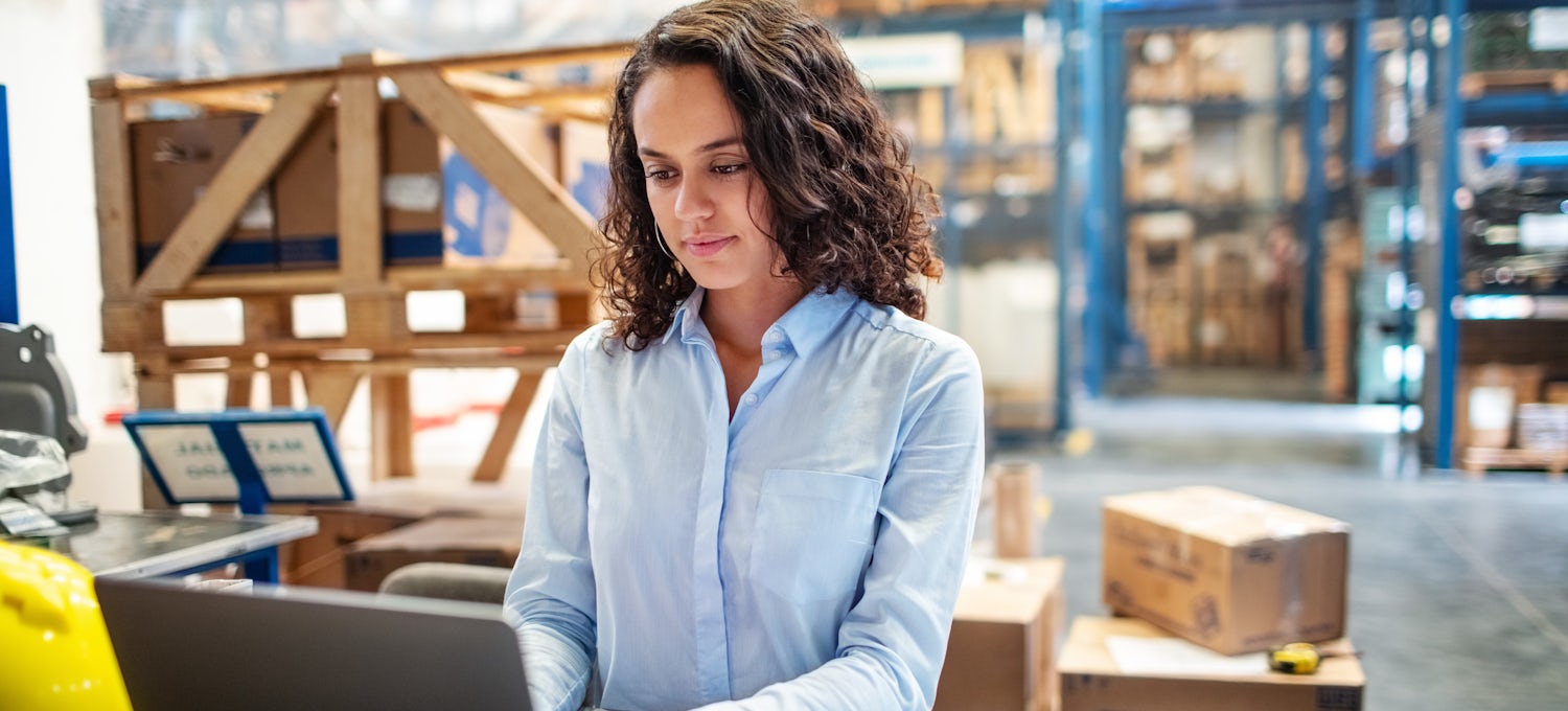 [Featured image] A businesswoman sits at a laptop in a warehouse and uses the SCOR model to improve her organization's supply chain management decisions. 
