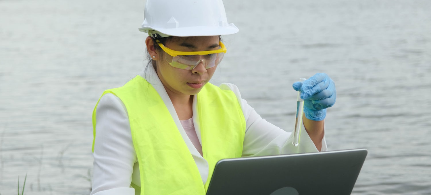 [Featured Image] An ecologist holes a vial of water and a laptop while working in the field using systems thinking skills. 
