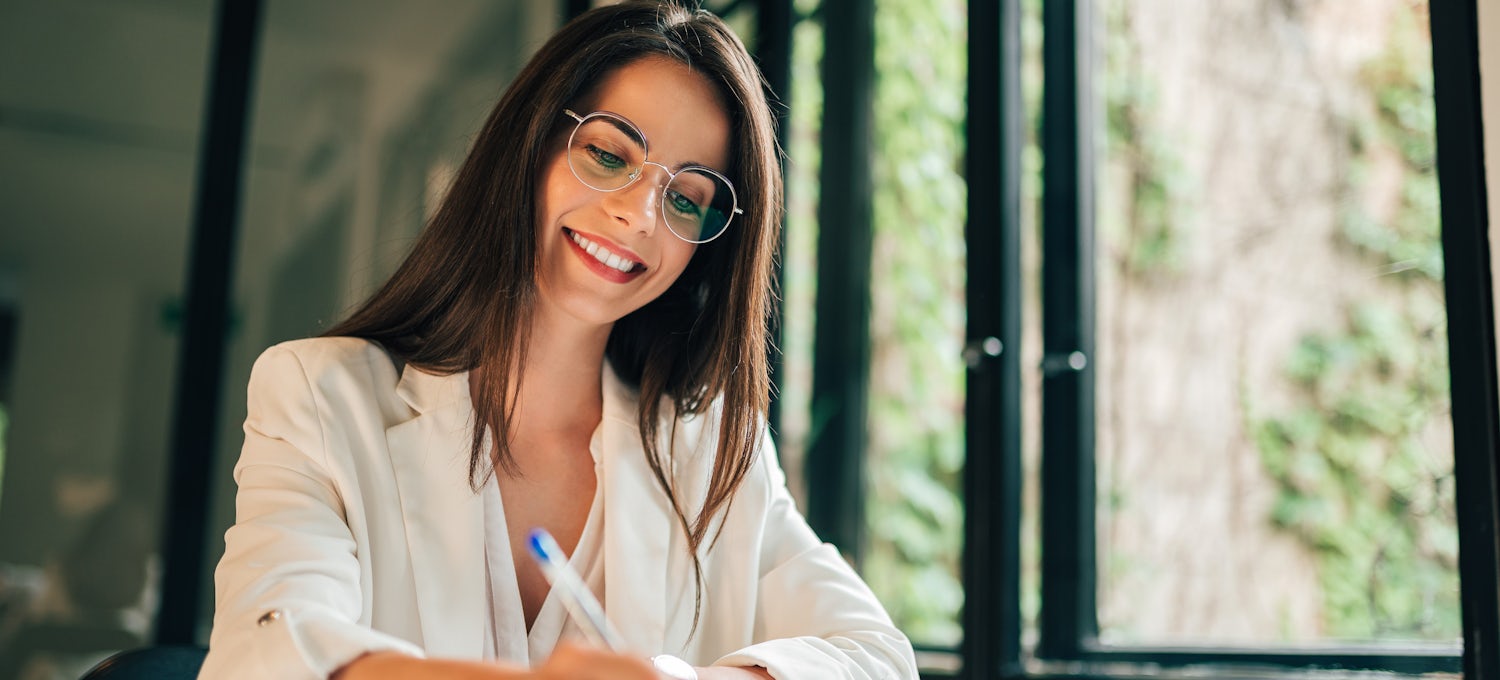 [Featured image] Job applicant wearing a white blazer and glasses works on creating an effective cover letter next to some windows.