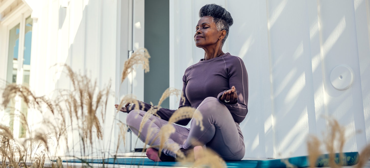 [Featured image] A fitness instructor wearing purple workout clothes sits outside on a blue yoga mat in a meditation pose.