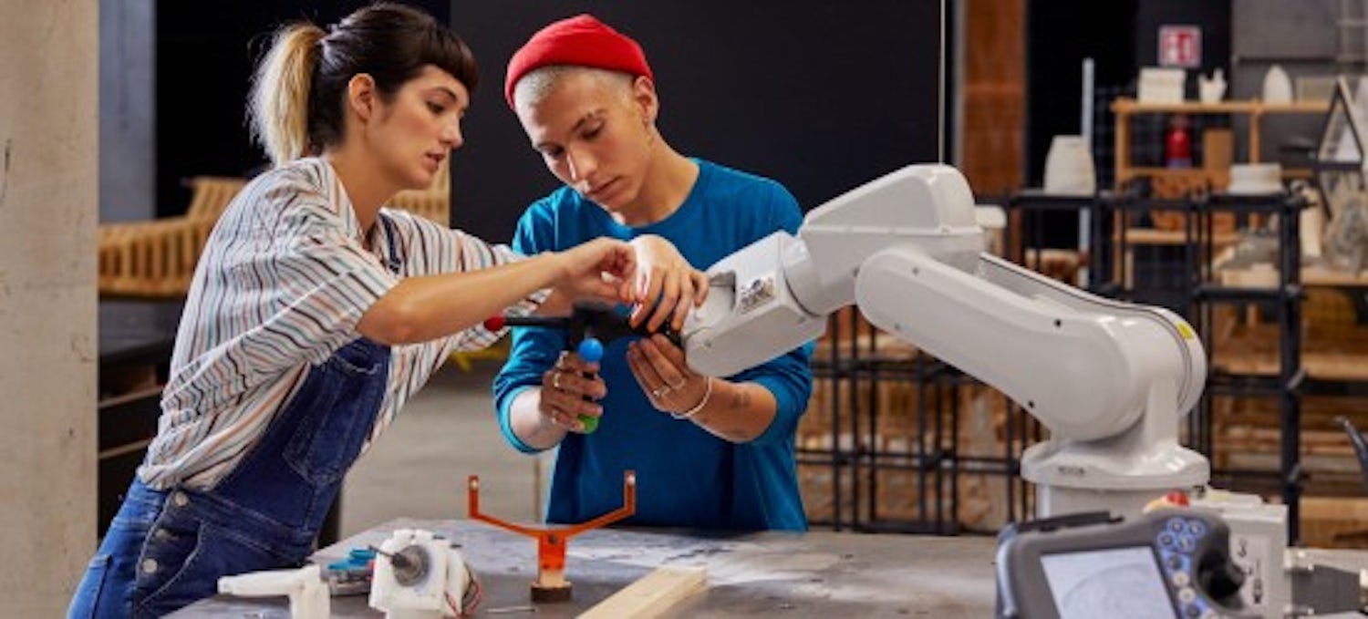 [Featured Image] Two robotics engineers work together on a robotic arm in a warehouse.